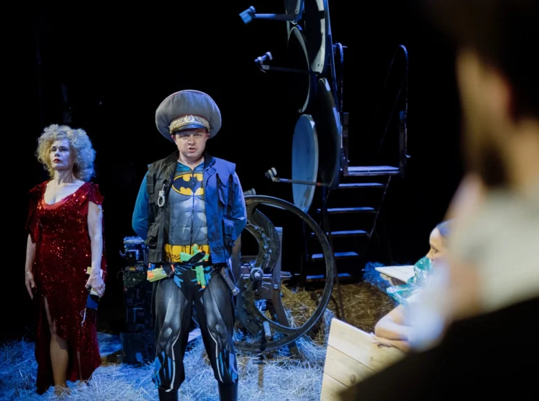 Spektakl teatralny. Na scenie stoją obok siebie kobieta w czerwonej, błyszczącej sukni i mężczyzna w stroju supermana, z czapką od nazistowskiego munduru. Obok nich scenografia przedstawiająca fragment jakiejś maszyny, być może rolniczej.