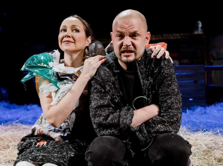 Spektakl teatralny. Na pierwszym planie na scenie siedzą obok siebie kobieta i mężczyzna. Kobieta obejmuje mężczyzną. Za nimi leży słoma. Z tyłu czarne tło.