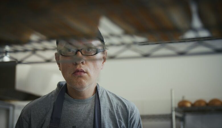 Kadr z filmu. Wnętrze piekarni. Zbliżenie na twarz i ramiona mężczyzny w kuchennym fartuchu. Mężczyzna patrzy na leżące na półce bochenki chleba.