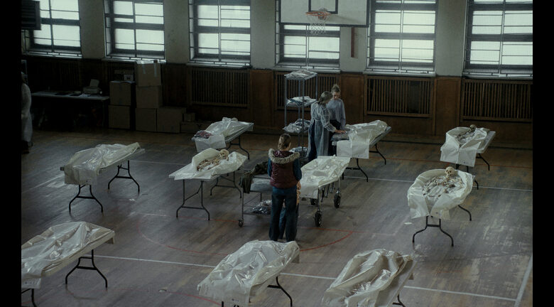 Kadr z filmu. W szkolnej sali gimnastycznej na stojących w rzędach stołach leżą ludzkie szkielety. Przy stołach stoją trzy kobiety. Jedn z nich ubrana jest w fartuch jednorazowy.