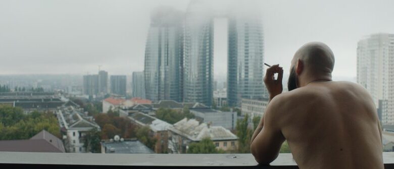Kadr z filmu. Na pierwszym planie zbliżenie na pokazanego od tyłu mężczyznę z nagim torsem. Mężczyzna opiera się o parapet okna lub balustradę balkonu i pali papierosa. Przed nim rozciąga się szeroki widok z góry na miasto.