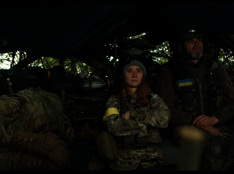 Kadr z filmu. Obok siebie stoją kobieta i mężczyzna w ukraińskich mundurach żołnierskich. Obok nich leży i śpi jeszcze jedna osoba w mundurze. Nad nimi widoczne niskie zadaszenie, przez otwory w zadaszeniu widać zielone drzewa.