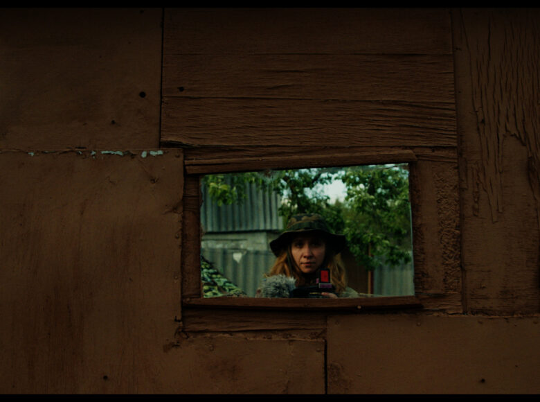 Kadr z filmu. Zbliżenie na ścianę. Pośrodku ściany znajduje się otwór przez który widać twarz kobiety w kapeluszu z tkaniny we wzór moro. Za kobietą widzimy ogrodzenie i drzewa.