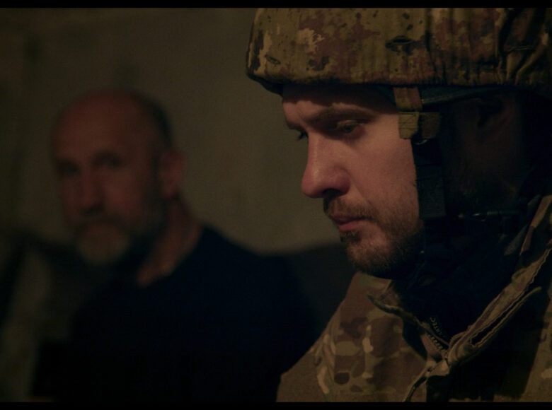 Kadr z filmu. Półmroczne pomieszczenie. Na pierwszym planie zbliżenie na twarz i ramiona młodego mężczyzny w mundurze żołnierza. W tle siedzi drugi, starszy mężczyzna w cywilnym ubraniu.