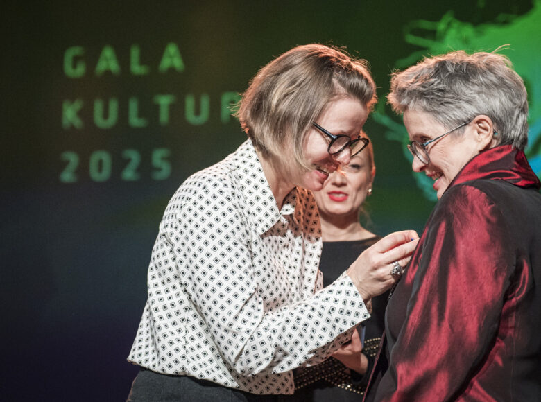 Uroczystość na scenie. Elegancko ubrana kobieta przypina medal Annie Nawrot, założycielce Galerii Białej w Lublinie.
