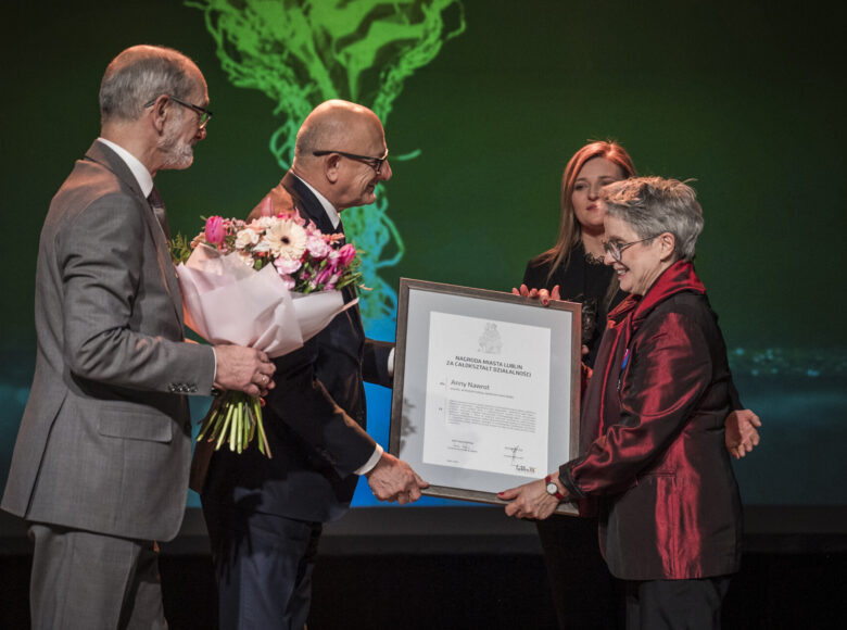 Uroczystość na scenie. Anna Nawrot, założycielka Galerii Białej w Lublinie, otrzymuje dyplom od prezydenta Lublina, Krzysztofa Żuka. Obok stoi zastępczyni prezydenta Beata Stepaniuk-Kuśmierzak oraz lubelski radny Jarosław Pakuła, który trzyma w rękach bukiet kwiatów.