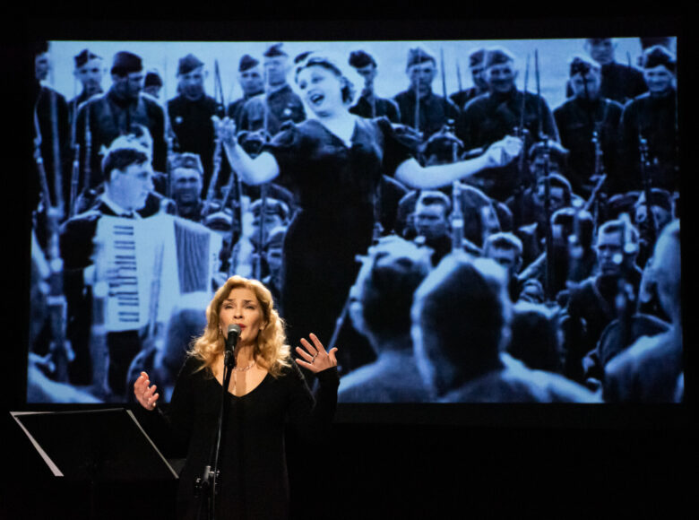 Spektakl teatralny. Na scenie stoi kobieta w czarnej sukni, która mówi lub śpiewa do mikrofonu. Za nią na ekranie wyświetlana jest scena z dwudziestego wieku przedstawiająca kobietę w eleganckiej sukni, śpiewającą dla otaczających ją żołnierzy.