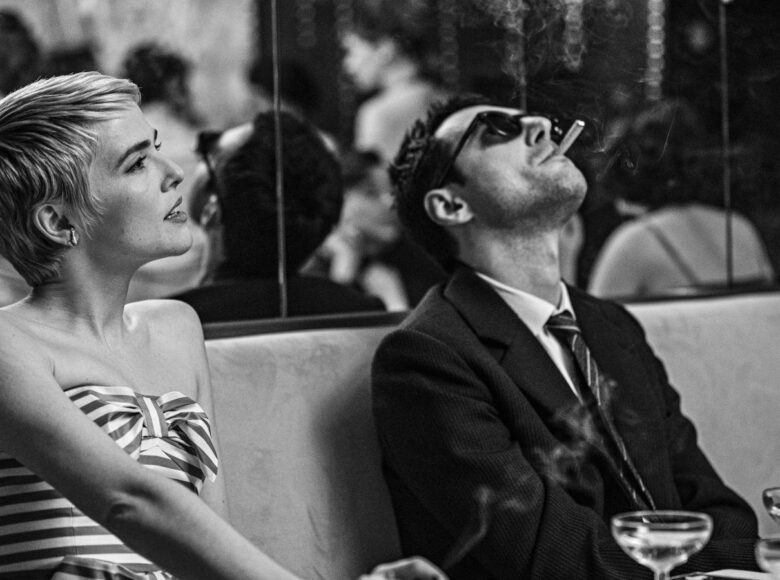 Kadr z filmu czarno biały. Kobieta i mężczyzna elegancko ubrani siedzą przy stoliku w restauracji. Oboje palą papierosy.