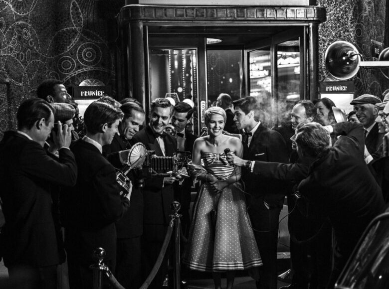 Fotografia czarno biała. Przed wejściem do eleganckiego budynku stoją elegancko ubrani kobieta i mężczyzna. Wokół nich duża grupa reporterów w garniturach i z aparatami fotograficznymi w dłoniach.