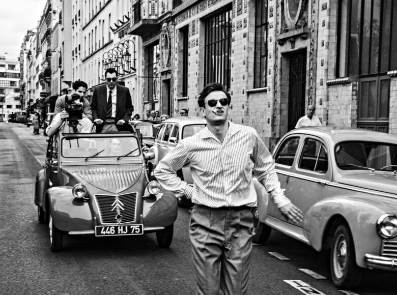 Kadr z filmu czarno biały. Miejska ulica, lata pięćdziesiąte lub sześćdziesiąte dwudziestego wieku. Na pierwszym planie, wśród samochodów spaceruje mężczyzna z papierosem w ustach. Jest filmowany przez mężczyzn, którzy jadą za nim samochodem. Wzdłuż ulicy stoją stare, eleganckie kamienice. Pod kamienicami stoją ludzie obserwujący scenę filmowania mężczyzny.