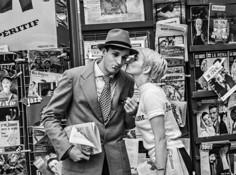 Kadr z filmu czarno białego. Miejska ulica, lata sześćdziesiąte dwudziestego wieku. Na tle kiosku z gazetami stoją obok siebie młoda dziewczyna i młody chłopak. Dziewczyna całuje chłopaka w policzek.