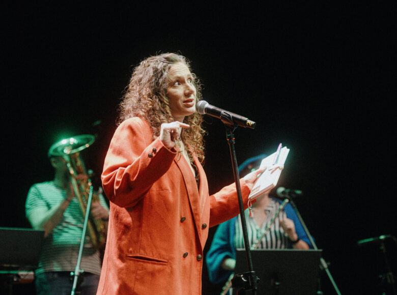 Na scenie kobieta przed mikrofonem. Za nią kobieta i mężczyzna grający na instrumentach muzycznych. Za nimi czarne tło.