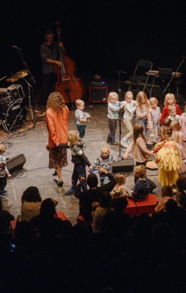 Pośrodku dużej sali grupa tańczących dzieci. Z boku stoi kilka dorosłych osób, grających na instrumentach muzycznych.