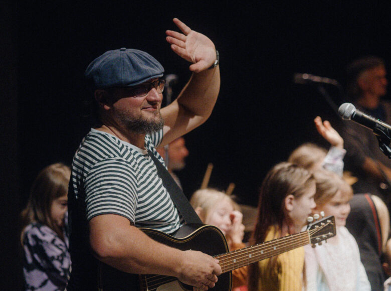 Na pierwszym planie mężczyznak, ktory w jednej ręce trzyma gitarę, a drugą rekę unosi do góry w geście pozdrowienia. Za mężczyzną stoi kilkoro dzieci, także machających rękami. Przed dziećmi stoi kilka mikrofonów. Na ostatnim planie czarne tło.