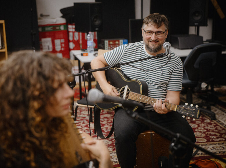 W pomieszczeniu przed mikrofonami siedzą kobieta i mężczyzna. Mężczyzna gra na gitarze, kobieta gra na cytrze.