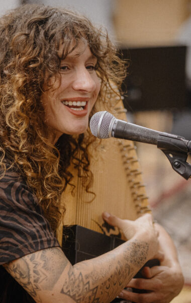 Kobieta siedzi na krześle przed mikrofonem. W ręku trzyma cytrę i śpiewa do mikrofonu.