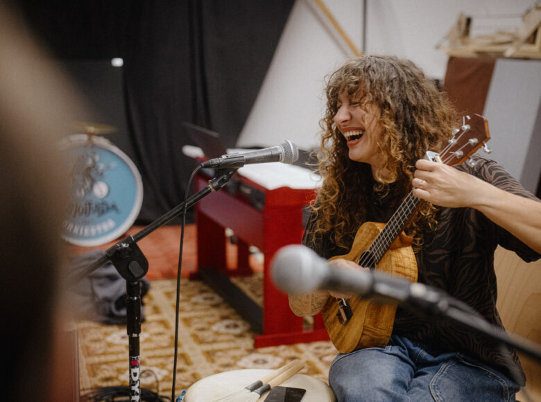 W pomieszczeniu przed mikrofonem siedzi na krześle kobieta i gra na gitarze.