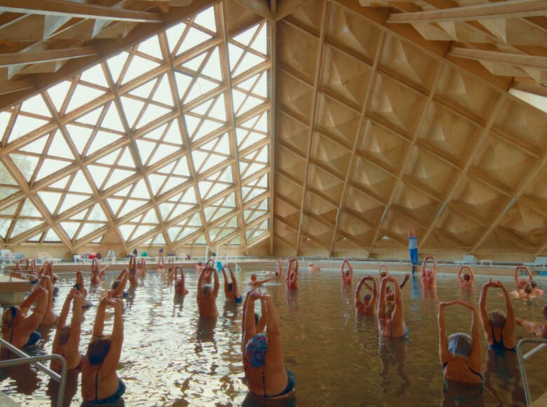 Kadr z filmu. W krytym basenie stoi duża grupa starszych kobiet wykonujących ćwiczenia gimnastyczne.