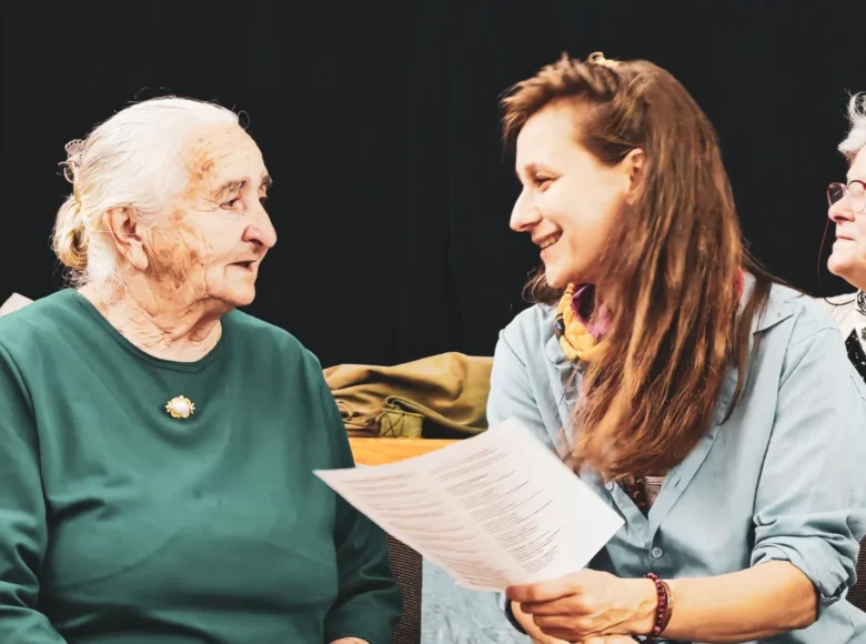 W pomieszczeniu siedzą obok siebie rozmawiając dwie kobiety, stara i młoda. Za nimi siedzi jeszcze jedna starsza kobieta. Z tyłu czarne tło.