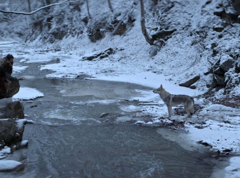 Kadr z filmu. Zimowy krajobraz. Nad strumieniem siedzi mężczyzna, mówiący do psa, który stoi na pobliskim drugim brzegu. Dookoła śnieg i drzewa.