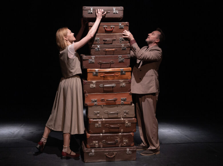 Przedstawienie teatralne. Na scenie kilkanaście staroświeckich walizek podróżnych, ułożonych jedna na drugiej w wysoki stos. Po obu stronach stosu walizek stoją kobieta i mężczyzna, kładąc na stos kolejną walizkę. Z tyłu czarne tło.