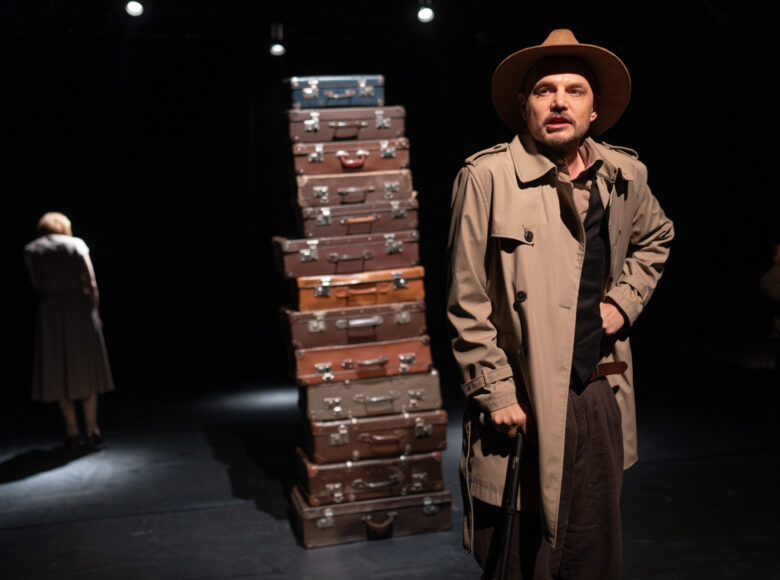 Spektakl teatralny. Na środku sceny kilkanaście staroświeckich walizek podróżnych, ułożonych jedna na drugiej. Przed nimi stoi mężczyzna w eleganckim płaszczu i kapeluszu. Na dalszym planie po dwóch bokach sceny widzimy dwie osoby: z jednego boku sceny kobietę stojącą plecami do widowni, a z drugiego boku sceny siedzącego mężczyznę. Z tyłu ciemne, czarne tło.