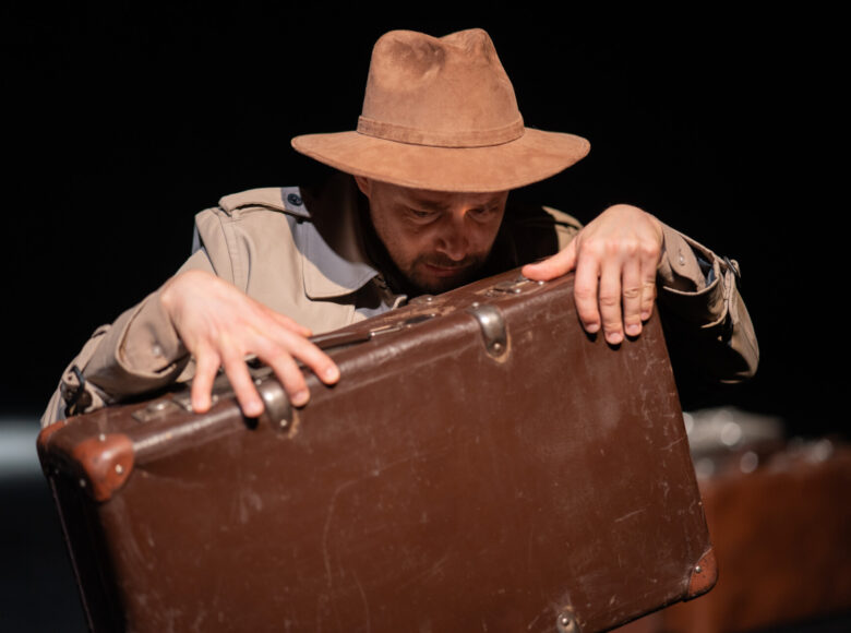 Przedstawienie teatralne. Zbliżenie na mężczyznę w kapeluszu, trzymającego starą walizkę podróżną.