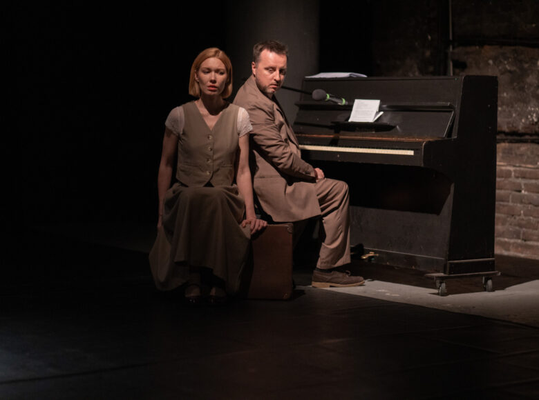 Przedstawienie teatralne. Na scenie stoi pianino, przy nim na starej walizce siedzą mężczyzna i kobieta.