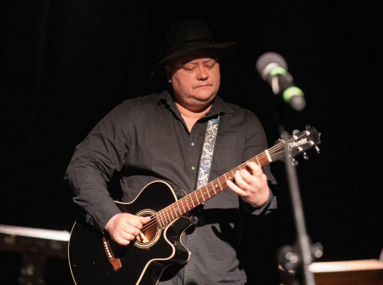 Spektakl teatralny. Przed mikrofonem stoi mężczyzna grający na gitarze. Za nim czerne tło.