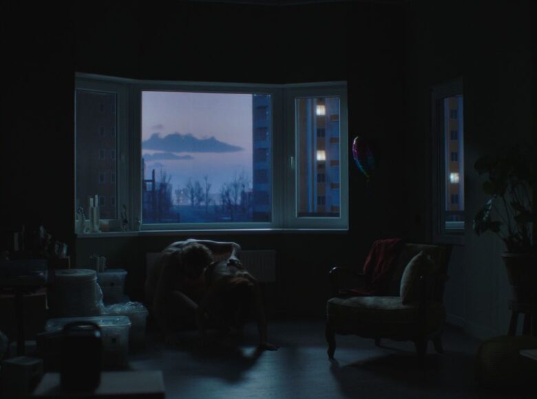 Kadr z filmu. Pokój w mieszkaniu, półmrok. Pod otwartym oknem na podłodze klęczą dwie osoby uprawiające seks. Przez okno widać bloki mieszkalne, drzewa i ściemniające się, wieczorne niebo.