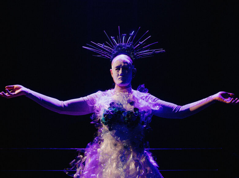 Na scenie, na czarnym, ciemnym tle stoi kobieta z rozpostartymi szeroko ramionami. Na twarzy ma maskę. Ubrana jest w suknię wykonaną z pociętych plastikowych butelek na wodę mineralną. Na głowie nosi ozdobę która może kojarzyć się z koroną amerykańskiej Statuy Wolności lub z aureolami świętych na starych obrazach.