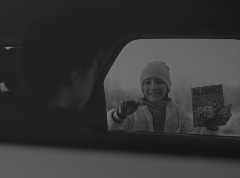 Kadr z filmu czarno biały. Wnętrze samochodu osobowego, w którym siedzi chłopiec. Za oknem samochodu stoi uśmiechnięta dziewczynka i pokazuje chłopcu książkę z komiksowym rysunkiem na okładce.