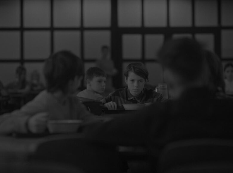 Kadr z filmu czarno biały. W szkolnej stołówce dzieci jedzą posiłek. W centrum kadru siedzi chłopiec, na którego patrzą inne dzieci siedzące dookoła niego.