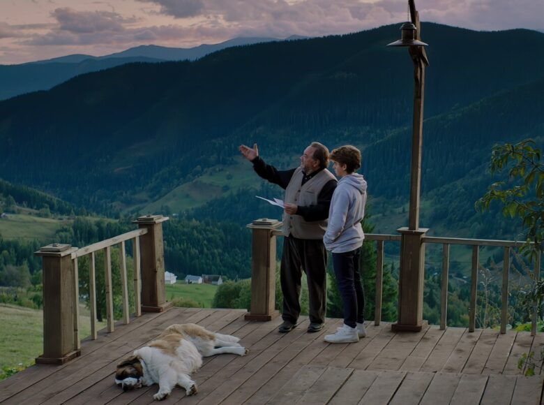 Kadr z filmu. Na tarasie drewnianego, wiejskiego domu stoją starszy mężczyzna i młody chłopak. Patrzą na widoczne dookoła,zalesione góry. Obok nich leży odpoczywający owczarek.
