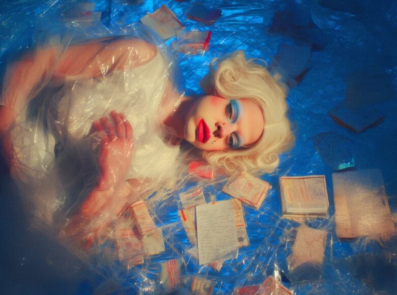 Młoda dziewczyna w mocnym makijażu i peruce blond leży na niebieskiej podłodze. Wokół niej leżą papierowe formularze i druki. Dziewczyna, podłoga i papiery przykryte są przezroczystą folią.