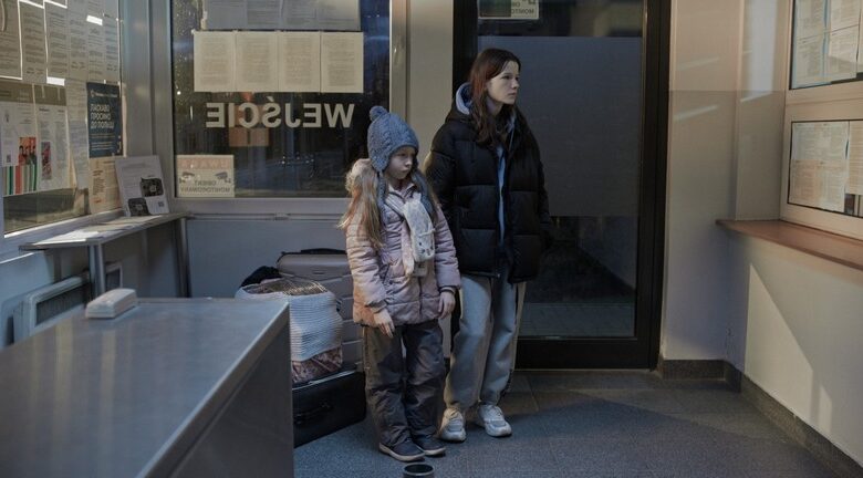 Kadr z filmu. W pomieszczeniu użyteczności publicznej obok okienka recepcji stoi młoda dziewczyna z kilkuletnią dziewczynką. Obie ubrane są w zimowe ubrania. Obok nich stoją bagaże.