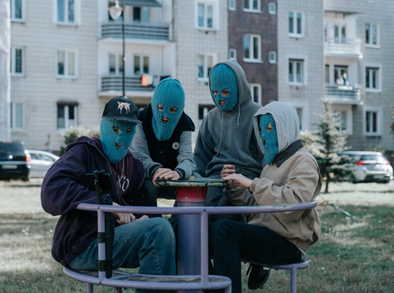 Kadr z filmu. Na pierwszym planie karuzela na placu zabaw. Na karuzeli siedzi czterech chłopaków w dresowych bluzach, dżinsowych spodniach i w niebieskich, włóczkowych kominiarkach na twarzach. W tle trawnik, blok mieszkalny i samochody zaparkowane pod blokiem.