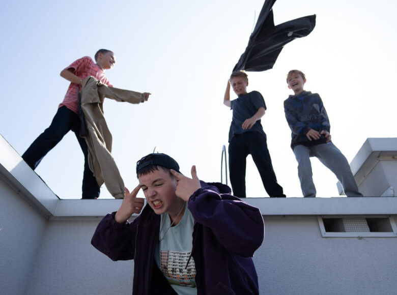 Kadr z filmu. Na dachu budynku stoi czterech dziesięcio lub kilkunastoletnich chłopców. Ubrani są w luźne, sportowe ubrania. W tle jasne, bezchmurne niebo.