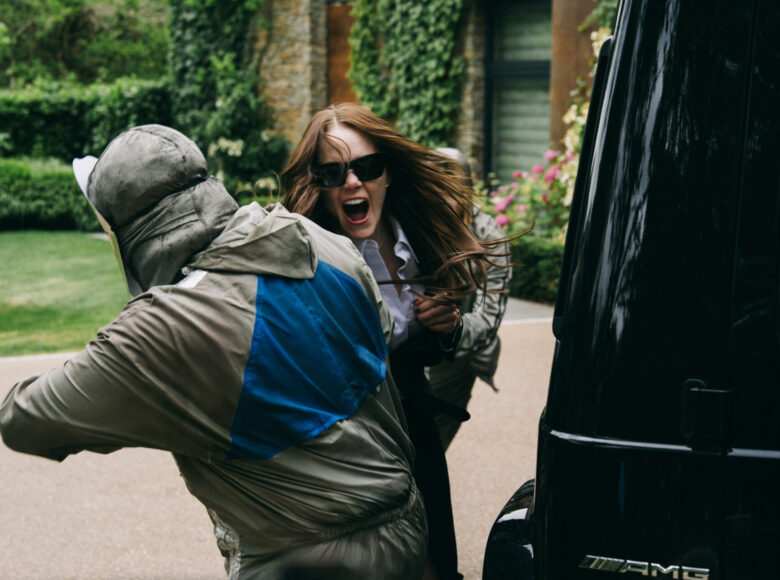 Kadr z filmu. Letni dzień. Na ulicy dwie osoby ubrane w kombinezony i maski ochronne trzymają wyrywającą się kobietę. Obok stoi duży, czarny samochód. W tle fragment domu, trawnik, krzaki i kwiaty.