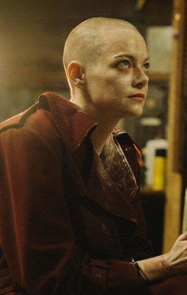 W pomieszczeniu, które wygląda jak składzik lub magazyn siedzi kobieta z ogoloną głową, ubrana w płaszcz. Na dalszym planie stoi silnie świecący reflektor.