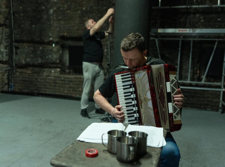 Przedstawienie teatralne. Na pierwszym planie przy małym stole siedzi mężczyzna i gra na akordeonie. Na dalszym planie kolumna, przy której stoi drugi mężczyzna i coś na niej wiesza. Za nim ceglasta ściana z małym okienkiem.