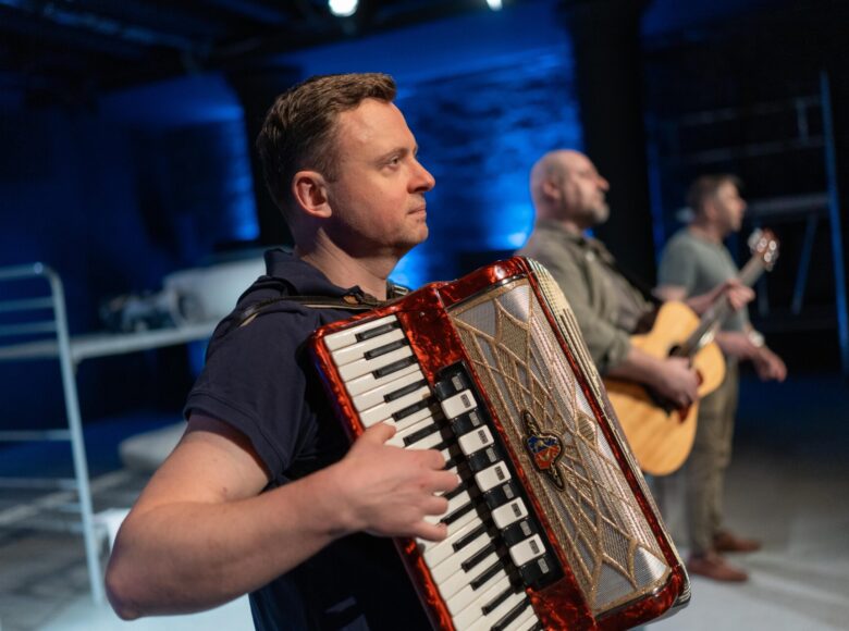 Przedstawienie teatralne. Na scenie stoją obok siebie trzej mężczyźni. Jeden gra na akordeonie, drugi na gitarze. Za nimi słabiej oświetlone fragmenty scenografii.