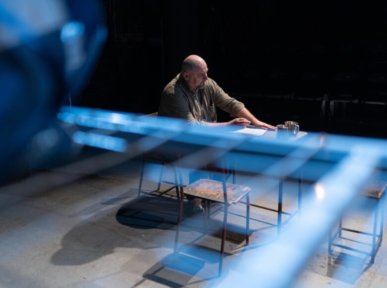 Przedstawienie teatralne. Zdjęcie zrobione od góry. Na scenie mężczyzna siedzący przy stole. Na stole przed mężczyzną leżą kartki i stoją dwa blaszane kubki. Przy stole stoją dodatkowe dwa puste krzesła.