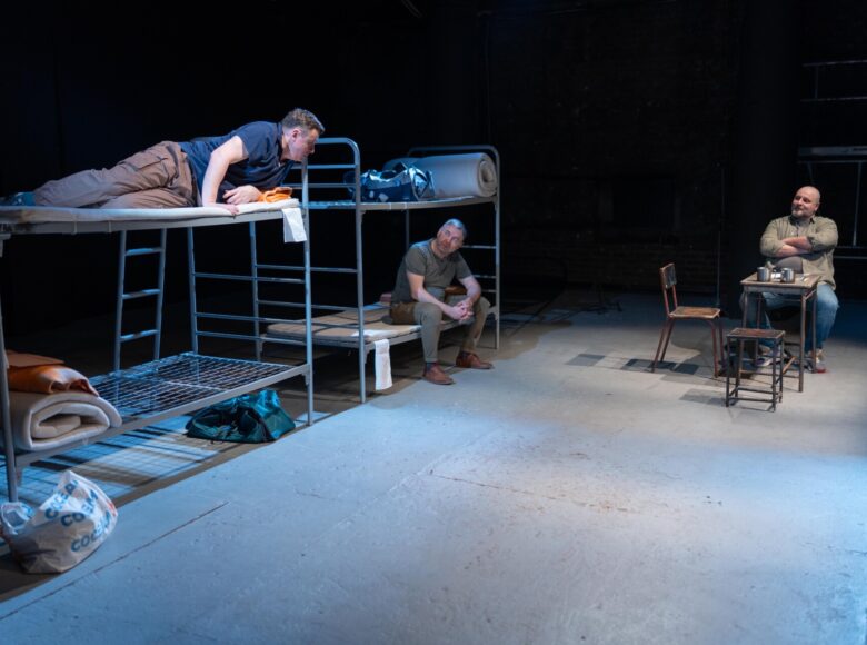 Spektakl teatralny. Na scenie stoją dwa łóżka piętrowe oraz stolik z trzema krzesłami. Na scenie widzimy także trzech mężczyzn, po jednym na każdym łóżku, a trzeciego siedzącego przy stole. Z tyłu ciemne tło, przedstawiające ścianę z małym okienkiem.