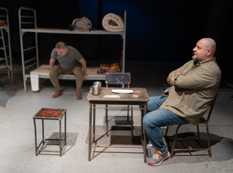 Spektakl teatralny. Na scenie na pierwszym planie przy stoliku siedzi mężczyzna. Na dalszym planie stoi łóżko piętrowe, na którym siedzi drugi mężczyzna. Na ostatnim planie czarne, ciemne tło.