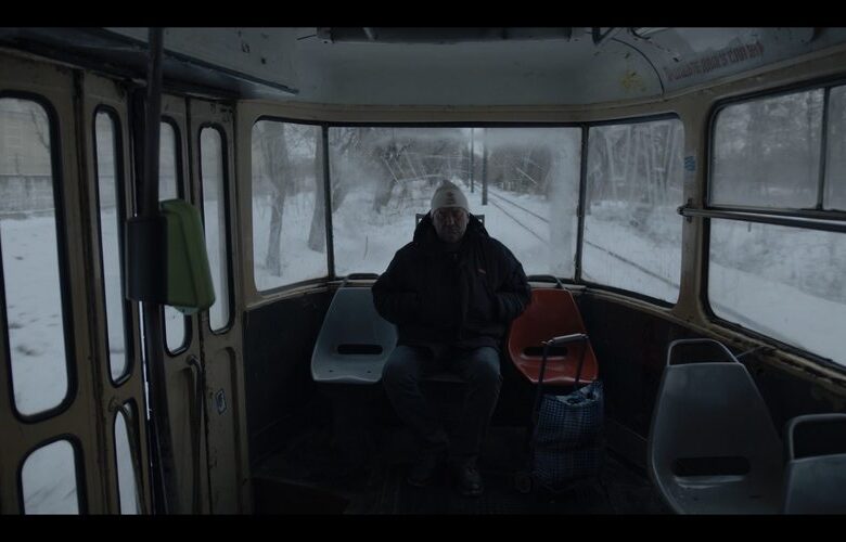 Kadr z filmu. We wnętrzu tramwaju siedzi mężczyzna w średnim wieku. Ubrany jest w zimowe ubrania. Za oknami tramwaju widać zaśnieżone drzewa i tory tramwajowe.