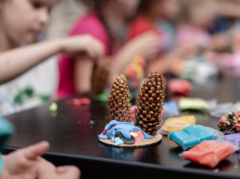 Zbliżenie na stojącą na stole kompozycję z dwóch podłużnych szyszek umocowanych na podstawce z plasteliny. W tle przy stole kilkoro dzieci wykonujących swoje kompozycje z plasteliny i szyszek.