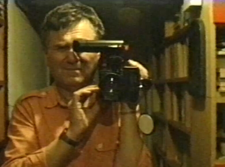 Na pierwszym planie mężczyzna w średnim wieku pokazany przodem. Mężczyzna filmuje coś trzymaną w rękach kamerą. Za plecami mężczyzny widać wnętrze mieszkania z półkami na książki.