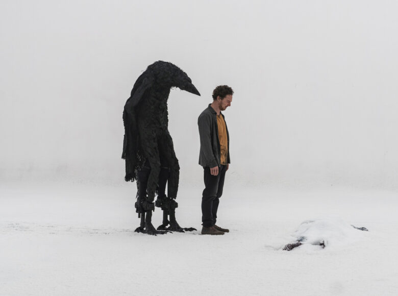 Kadr z filmu. Na śniegu, na tle białego nieba stoi mężczyzna oraz fantastyczna czarna postać z ciałem podobnym do ludzkiego i głową ptaka.