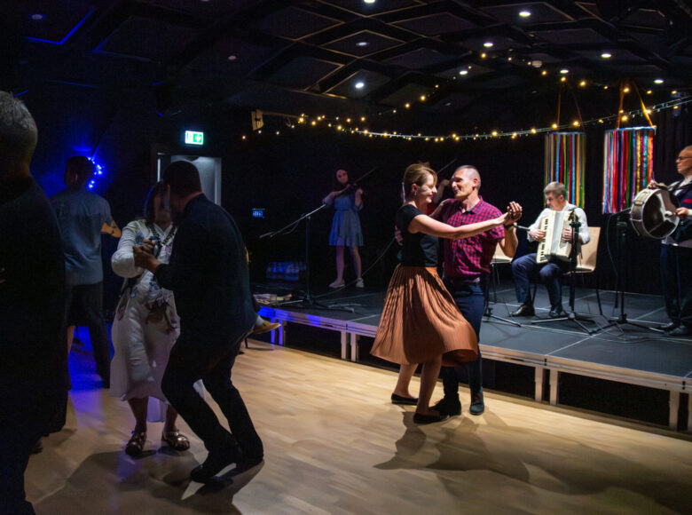 W sali ludzie tańczący w parach. Obok nich scena, na której widzimy osoby, przygrywające do tańca na skrzypcach, akordeonie i bębnie.